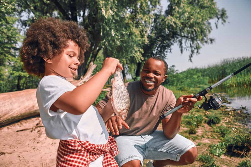   Слатки коврџави афроамерички дечак који гледа рибу у рукама док његов тата држи штап за пецање